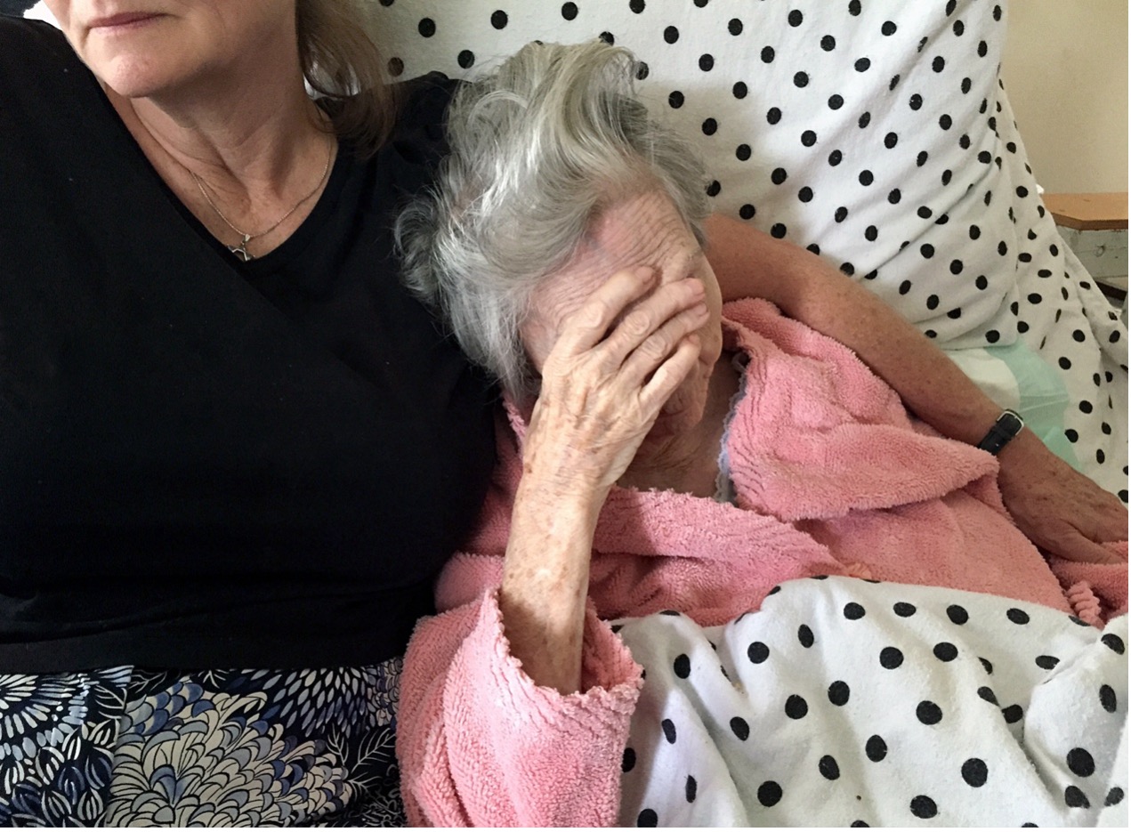 An elderly woman in a pink robe sits on a bed, covering her face with her hand. Beside her, another woman sits close, offering support. The bed is covered with a polka-dot blanket.