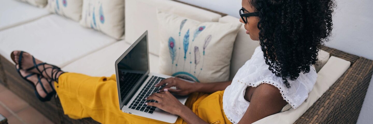 Woman working from home sitting on patio furniture while writing on her laptop