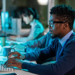 person in front of a computer with another person in background on computer